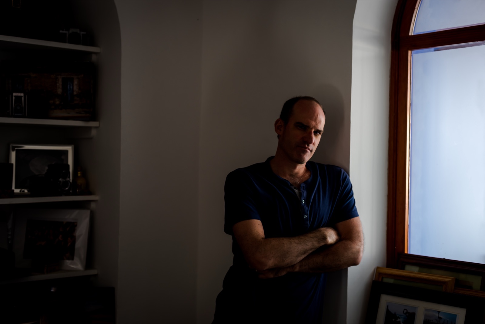 Douglas Ryan Favero standing with his arms crossed in a dim room beside a window